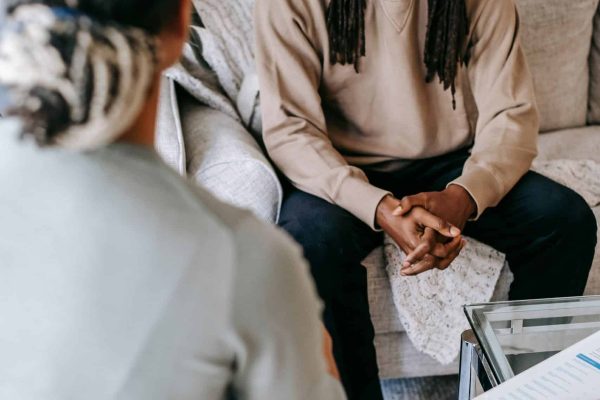 African American male wearing smart casual clothes sitting on sofa and talking to female psychologist or colleague in light studio
